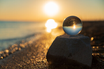 Glass lens globe at seaside on sandy beach on sea surf and blue sky background at sunset. Scenic seascape.