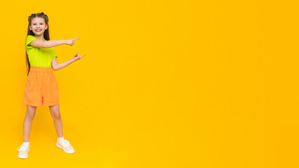 A child in shorts and a T-shirt. Happy childhood. A little girl with long hair enjoys the summer warmth inside and points to your advertisement on a yellow isolated background. Copy space. Banner.