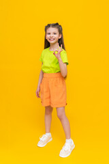 A child in shorts and a T-shirt. Happy childhood. A little girl in full height with long hair rejoices in the summer warmth inside and points to your advertisement on a yellow isolated background.