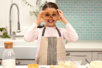 Cheerful european little girl in apron makes cookies, puts cookie cutters to eyes, has fun in...