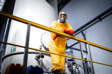 Portrait of factory worker in protective yellow suit, gas mask and gloves leaning against the fence inside chemicals production plant.