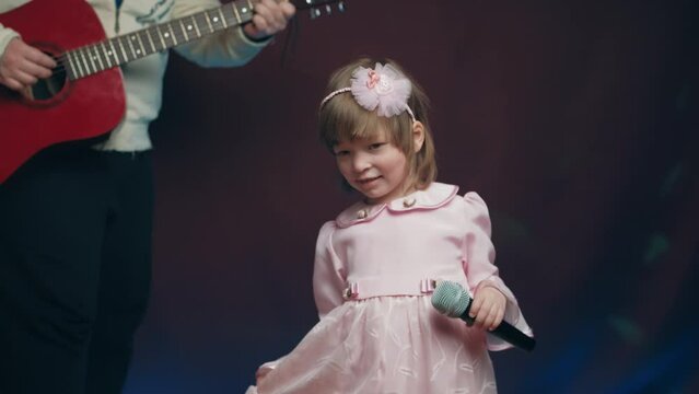 Little girl on stage in vintage dress, she sings into microphone and dances, her father plays acoustic guitar. Color music is shining. Performance on stage. Early development of children