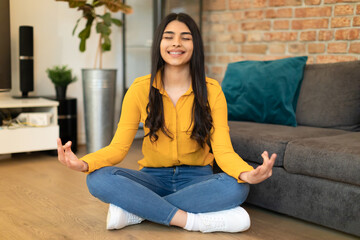 Cheerful spanish lady sitting on floor by couch, meditating with closed eyes and smiling at home, living room interior - obrazy, fototapety, plakaty