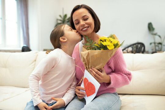 Cheerful little girl kissing european millennial woman with flowers hold postcard with heart in living room