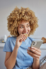 Pleased thoughtful curly haired woman eats cookie for breakfast uses mobile phone for searching morning news wears casual blue t shirt poses at kitchen focused aside. People and lifestyle concept