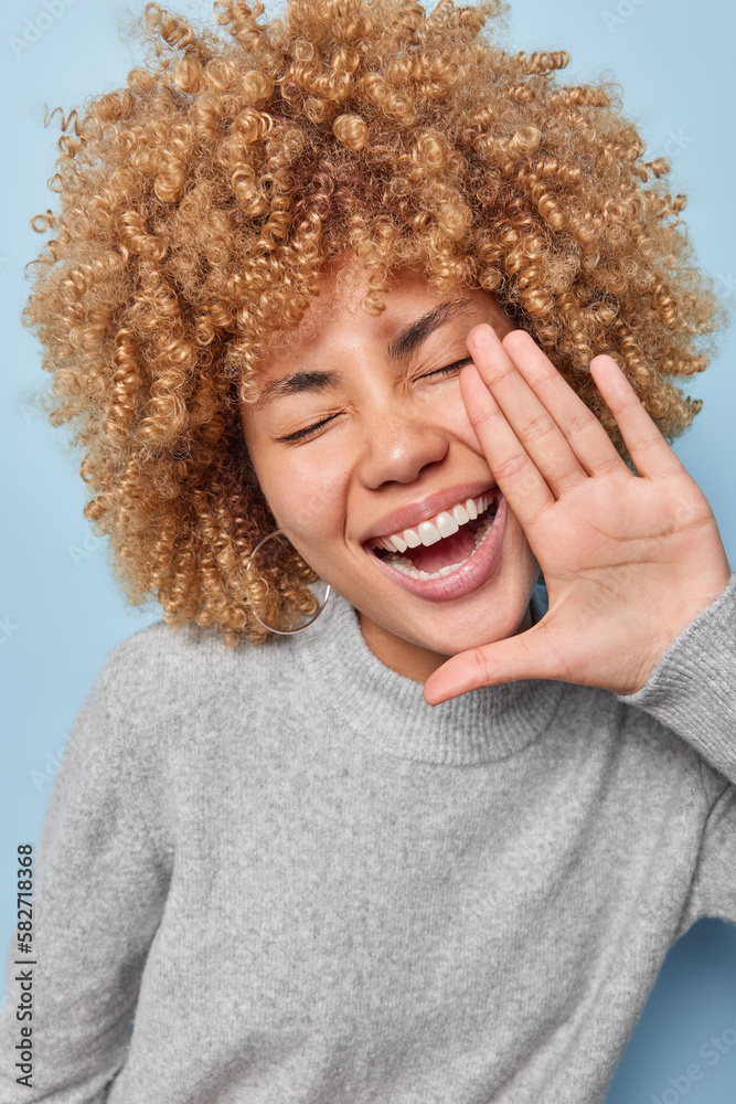 Wall mural vertical shot of cheerful curly haired adult woman keeps hand near opened mouth has fun dressed in g