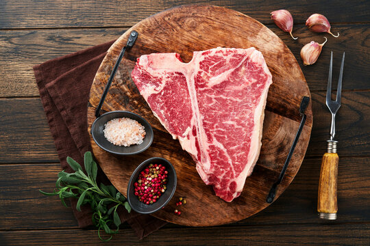 Thick Raw T-Bone Steak. Dry-aged Raw T-bone or porterhouse beef meat Steak on cutting boar with herbs and salt on dark background. Top view and copy space.