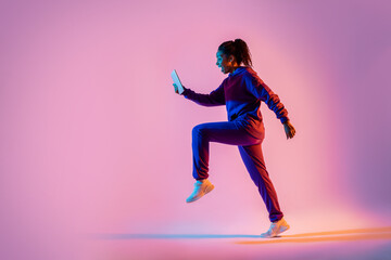Fototapeta na wymiar Full body length shot of black lady jumping or running, using cellphone over pink neon background, side view, copy space
