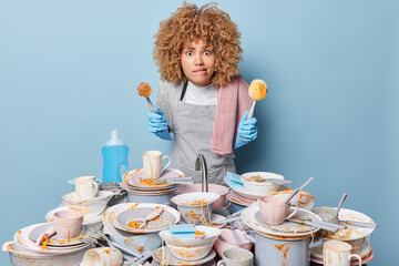 Worried busy housewife bites lips and looks at camera holds cleaning brush wears apron and rubber...