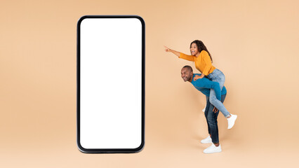 Great mobile app. Happy black couple presenting huge smartphone with mockup, man riding lady on back