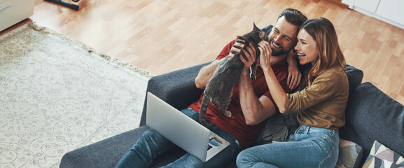 Happy young couple having fun with their cat while sitting on the couch at home together