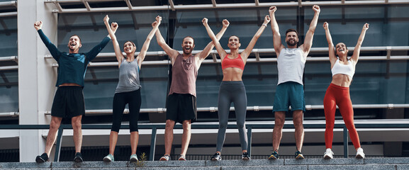 Group of happy young people in sports clothing keeping arms raised while standing outdoors © gstockstudio