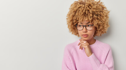 Portrait of curly haired woman has thoughtful face keeps hand under chin ponders on something wears transparent eyeglasses and pink knitted jumper isolated over white background blank space for text
