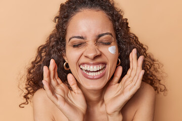 Fototapeta premium Daily skin care concept. Overjoyed curly haired woman applies facial cream for moisturising takes care of skin smiles toothily stands half naked isolated over brown background uses beauty product