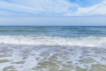 Blue cloudy sky above blue sea water, waves crashing sandy beach, windy weather. Summer vacation and holiday travel concept. Seascape nature background, beautiful seashore with water foam