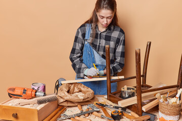 Professional female carpenter markes wood with pencil wears checkerd shirt denim overalls protective gloves poses in workshop uses different instruments for producing furniture isolated on beige wall