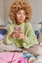 Vertical shot of pensive curly haired woman dressed in casual jumper and trousers holds mobile phone poses on bed with clothes around in messy bedroom. People home lifestyle and technology concept