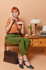 Unhappy sad woman with red hair has telephone talk keeps handset near ear looks displeased down has some troubles dressed in elegant old fashioned clothing sits on chair. Vintage style concept
