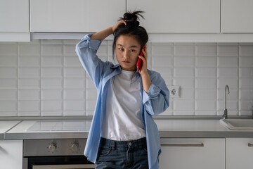 Upset dissatisfied Asian woman talking on phone arguing by mobile while standing in kitchen at...