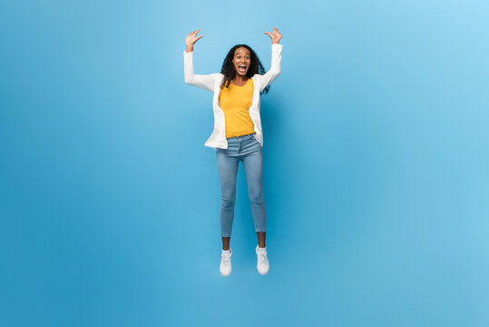 Full length portrait of smiling ecstatic African American woman jumping with arms raised in isolated light blue color studio background
