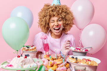 Happy curly haired woman smeared with cream after eating delicious sweet desserts winks eye looks gladfully at camera stands near festive table celebrates birthday surrounded by inflated balloons