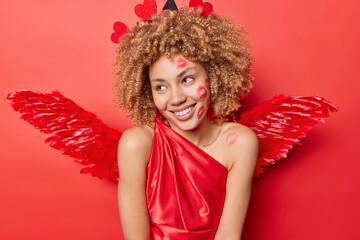 Pleased curly haired woman smiles gently has traces of kiss wears dress and wings has dreamy...