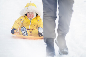Toddler baby rides in the snow on icesled, a winter playground. Mother woman sledding baby boy in yellow snowsuit. Kid age one year eight months