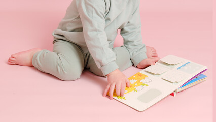 Happy toddler baby is reading a book on studio pink background. Child boy plays with a book. Kid...