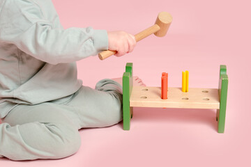 Happy toddler baby plays with a hammer and wooden nails on a studio pink background. Child boy with educational toy. Kid age one year eight months