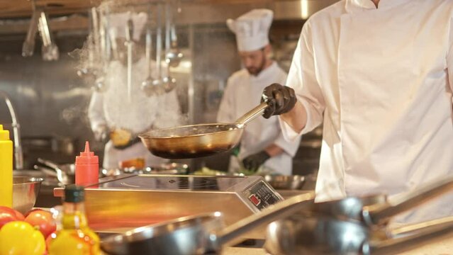 Close-up portrait of delicious food being frying on pan and mixed by handsome young caucasian professional chef in white uniform. Attractive cook cooking tasty dishes in modern kitchen.