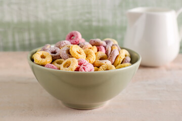 Bowl of colorful cereal rings and pitcher with milk on white wooden table near green wall