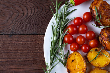 Part of a plate with a dish of chicken, potatoes, cherry tomatoes and rosemary on a wooden table.