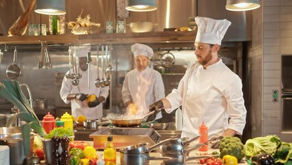 Portrait of beautiful professional caucasian chef cooking vegetables frying on pan and mixing. Attractive skillful bearded male cook working at modern hotel kitchen smiling enjoying work. - Powered by Adobe
