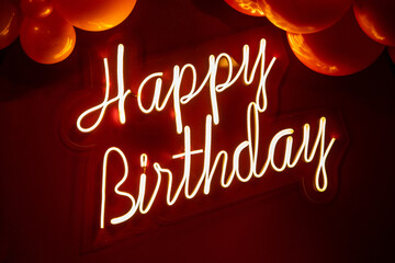 Escrito "Feliz Aniversário" com luz de neon sobre fundo vermelho e com vários balões ao redor.