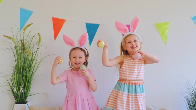 Two happy little girls sisters in rabbit ears jumping and dancing on the couch holding easter eggs.