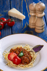 spaghetti bolognese pasta in a white plate, salt, pepper, cheese and tomatoes on blue wooden background