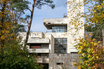 A light concrete building in the form of cubism with a view through colorful autumn trees and a...