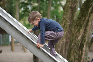 Little Boy trying to going up the slider. Preschool child playing on outdoor playground. Happy...