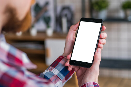 Mockup image - over shoulder view: man entrepreneur hands holding black smartphone with white blank screen in home interior. Mock up, copyspace, template, entertainment and technology concept