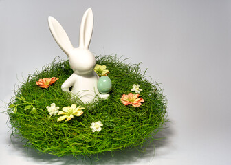 Porzellan Osterhase hält ein grünes Osterei, in einem Gras Nest mit Blumen Hintergrund grau,...