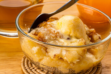 Closeup view of delicious homemade peach cobbler with a scoop of vanilla ice cream and metal spoon in transparent glass bowl on bamboo mat with a glass of fresh orange juice and a cup of hot tea.