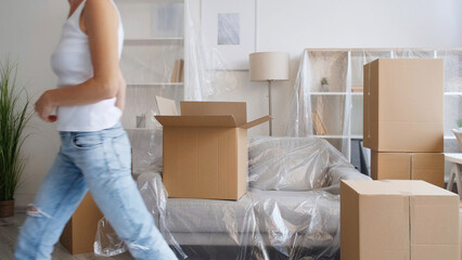 Fototapeta na wymiar Home moving. Flat relocation. Removal service. Unrecognizable woman leaving room with carton boxes in modern apartment with polyethylene covered furniture.