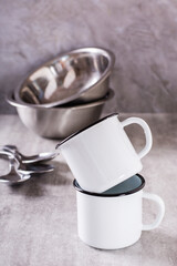 Two white empty metal mugs and metal bowls and spoons on a gray vertical background