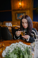 The girl with dark hair is drinking tea and tapping on her phone at a morning café at a table where a beautiful bouquet of white flowers is lying