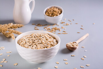 Raw oatmeal in a bowl with a wooden spoon and ears on a gray background.