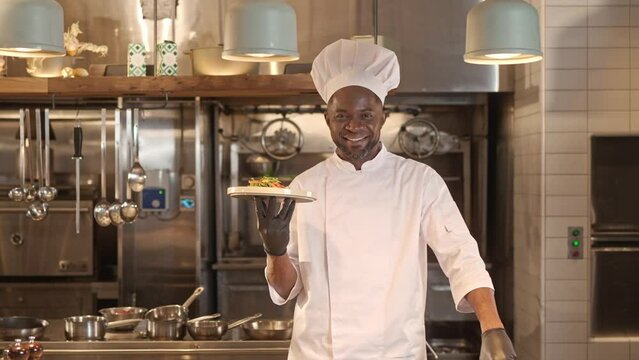 Portrait of professional happy male African American chef serving and presenting delicious salad looking at camera smiling. Attractive successful cook working at kitchen of fancy restaurant.