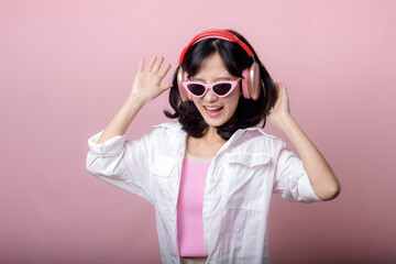 happy young asian woman model with stylish trendy sun glasses enjoy listening music by headphone audio and dancing isolated on pink studio background. technology, girl fashion, accessory concept.