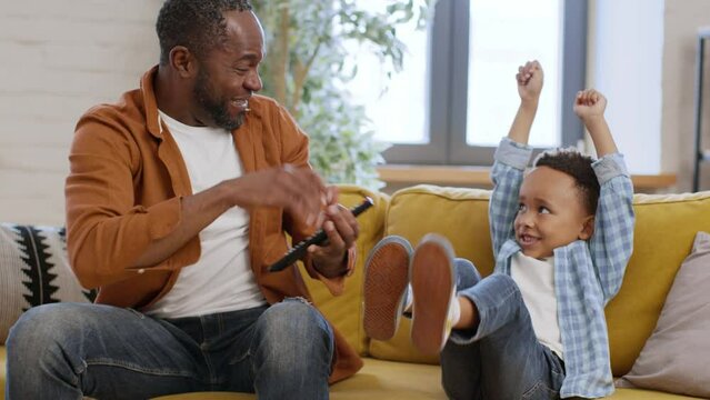 Emotional little african american boy watching game on tv with dad at home, celebrating goal, tracking shot, slow motion
