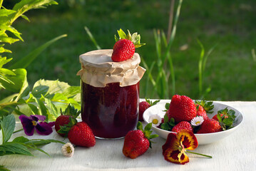 Homemade strawberry jam in glass jar and ripe fresh berries red strawberry with flowers daise and pansy, green leaves on a white wooden table on green natural blur background