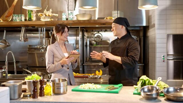 Portrait of successful beautiful female manager of restaurant using tablet and talking with professional asian male chef chopping vegetables at kitchen. Two attractive colleagues working together.